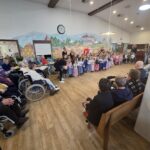 Več oseb sedi ob stenah prostora, medtem ko skupina otrok stoji v vrsti na sredini in izvaja nastop.