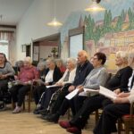 Skupina oseb sedi v vrsti na stolih in drži liste papirja, medtem ko sodelujejo v skupni dejavnosti.