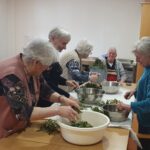 Skupina oseb stoji okoli mize in čisti zelenje ter ga odlaga v sklede.