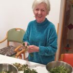 Ženska stoji ob mizi in drži šop zelenja, na mizi so sklede z očiščenimi listi.