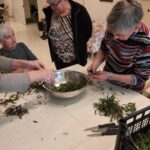 Več oseb stoji okoli mize in pripravlja regrat, ki ga dajejo v skledo.