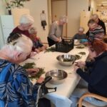 Skupina starejših sedi in stoji ob mizi ter skupaj čisti in pripravlja regrat.