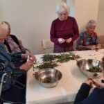 Več oseb sedi okoli mize in s pomočjo nožev pripravlja regrat za solato.