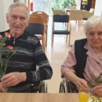 Moški in ženska sedita za mizo na invalidskih vozičkih in v rokah držita nageljna.
