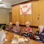 Skupina oseb sedi za mizo in ploska v prostoru z omarami; na steni je napis »Vse najboljše!«.