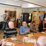 Oseba igra harmoniko, medtem ko druge osebe sedijo za mizo in ploskajo.