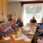 Osebe sedijo za mizo, držijo kozarce in nazdravljajo; na mizi je torta.