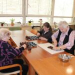 Tri osebe sedijo za dolgo leseno mizo v kuhinjskem prostoru z velikimi okni in pripravljajo čokoladne sladice. Ena oseba drži čokolado in jo lomi, druga oseba reže čokolado na deski, tretja oseba drži pekač z oblikovanimi kosi čokolade.