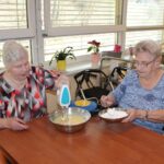 Dve osebi sedita za mizo v kuhinjskem prostoru in mešata testo. Ena oseba drži ročni mešalnik v posodi z rumenim testom, druga oseba drži žlico in dodaja moko iz kovinske posode.