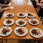 Na dolgi mizi stoji devet krožnikov s pripravljenimi čokoladnimi sladicami, okrašenimi z rdečo omako, vaniljevo kremo in čokoladnim prelivom v vzorcih. Okoli mize so vidne roke in deli teles oseb, ki sedijo.