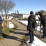 Oseba stoji ob ograji na prostem in govori proti kameri, medtem ko snemalna ekipa z video kamero in lučjo snema prizor.