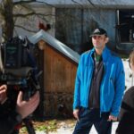 Moški stoji pred kamero na prostem, za njim je majhen lesen objekt, medtem ko snemalna ekipa pripravlja posnetek.