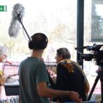 Snemalna ekipa z video kamero in mikrofonom snema osebo, ki stoji ob kletki z živaljo v notranjem prostoru ob oknu.