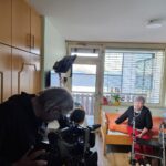 Snemalec snema žensko, ki sedi na postelji v sobi, ob njej stoji hodulja.