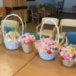 Na mizi so razporejene košarice z umetnimi cvetličnimi aranžmaji v pastelnih barvah.