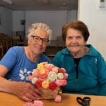 Dve ženski sedita za mizo in držita košarico z umetnimi cvetovi; na mizi so razporejeni posamezni cvetovi in škarje.