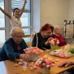 Več oseb sedi za mizo in izdeluje cvetlične aranžmaje iz umetnih cvetov; ena oseba stoji za njimi z dvignjenimi rokami.