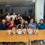 Skupina oseb sedi in stoji okoli mize z izdelanimi cvetličnimi košaricami ter gleda proti kameri.