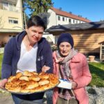 Dve ženski stojita na prostem; ena drži pladenj s pecivom, druga v rokah drži zavitek prtičkov.