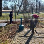 Dve osebi na prostem grabita suho listje ob ograji in drevesih.