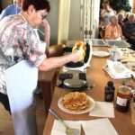 Ženska peče sveže vaflje na aparatu, na mizi pa so pripravljeni dodatki, kot so marmelada, čokolada in že pečeni vaflji.