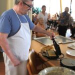 Moški sodeluje pri peki vafljev in preverja njihovo pripravljenost, medtem ko stanovalci v ozadju opazujejo dogajanje.