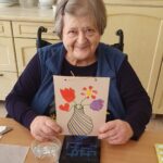 Ženska sedi za mizo in drži voščilnico z narisano vazo in cvetovi.