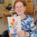 Ženska sedi na stolu in drži voščilnico z narisano vazo in cvetovi.