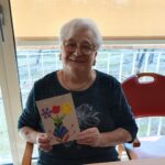 Ženska sedi za mizo in drži voščilnico z narisano vazo in cvetovi.