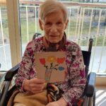 Ženska sedi na invalidskem vozičku in drži voščilnico z narisanimi cvetovi v vazi.