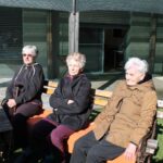 Tri osebe sedijo na klopi na prostem pred stavbo in počivajo na soncu.