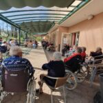 Več oseb sedi na stolih in invalidskih vozičkih pod nadstreškom ter sodeluje v skupinski aktivnosti na prostem.