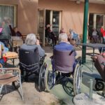 Več oseb, sedečih v krogu na stolih in invalidskih vozičkih, dviguje roke med skupinsko vadbo na terasi.