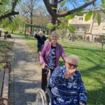 Tri osebe se po poti v parku premikajo z invalidskimi vozički; ena oseba hodi ob vozičku, druga oseba maha.