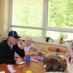 Več oseb sedi za mizo ob oknu, na mizi so skodelice, pecivo in ustvarjalni material.