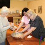 Več oseb stoji ob mizi in skupaj meša testo v kovinski posodi v skupnem prostoru.