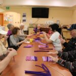 Več oseb sedi za dolgo mizo in izdeluje predmete iz vijoličnega ustvarjalnega materiala.