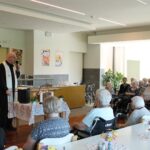 Duhovnik blagoslavlja velikonočne jedi, stanovalci pa zbrano spremljajo obred v skupnem prostoru.