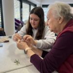 Mlajša udeleženka pomaga starejši gospe pri natančnem zavijanju jajca v tkanino z dodatkom naravnih materialov.