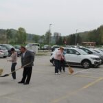 Štiri osebe na parkirišču pometajo tla z metlami in pobirajo smeti ob parkiranih avtomobilih.