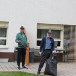 Dve osebi stojita ob stavbi; ena drži grablje, druga pa vrečo za odpadke.