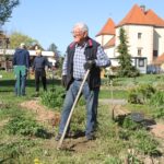 Oseba z lopato obdeluje zemljo v gredi, v ozadju druge osebe opravljajo vrtnarska dela.