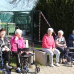 Več oseb sedi na klopi ob poti, pred seboj imajo hodulje, in spremlja dogajanje na prostem.