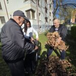 Tri osebe na travi pobirajo suho listje; ena oseba drži vrečo, druga prinaša kup listja.