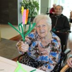 Starejša ženska na invalidskem vozičku nasmejana pokaže ročno izdelano barvito rožo.