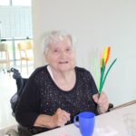 Starejša ženska sedi za mizo, drži izdelano rožo iz žičk in gleda proti fotografu.