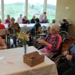 Skupina starejših oseb sedi za mizo in ustvarja rože iz barvnih materialov, medtem ko se v ozadju drugi pogovarjajo in opazujejo.