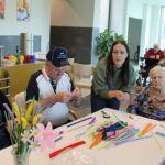 Starejši moški in ženska ob pomoči mlajše osebe izdelujejo rože iz pisanih žičk za mizo.