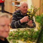 Oseba sedi za mizo in sestavlja šopek iz vejic in rumenih cvetov.