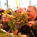 Oseba drži šopek iz zelenih vejic in rumenih cvetov, medtem ko druga oseba pomaga pri delu.